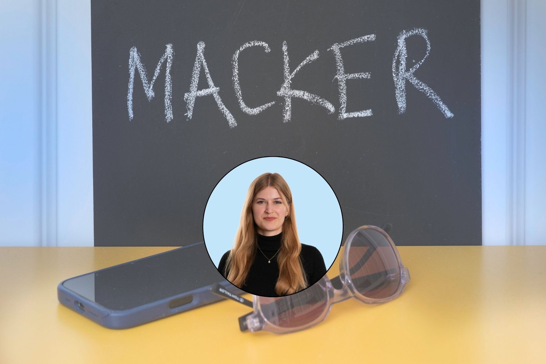 Kreidetafel mit Schriftzug "MACKER", Smartphone, Brille und Bild einer Frau auf Tisch.
