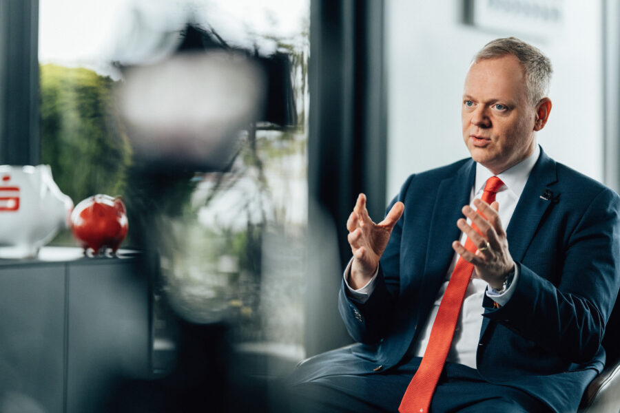 Ein Mann im Anzug spricht und gestikuliert. Im Hintergrund zwei Sparschweine.