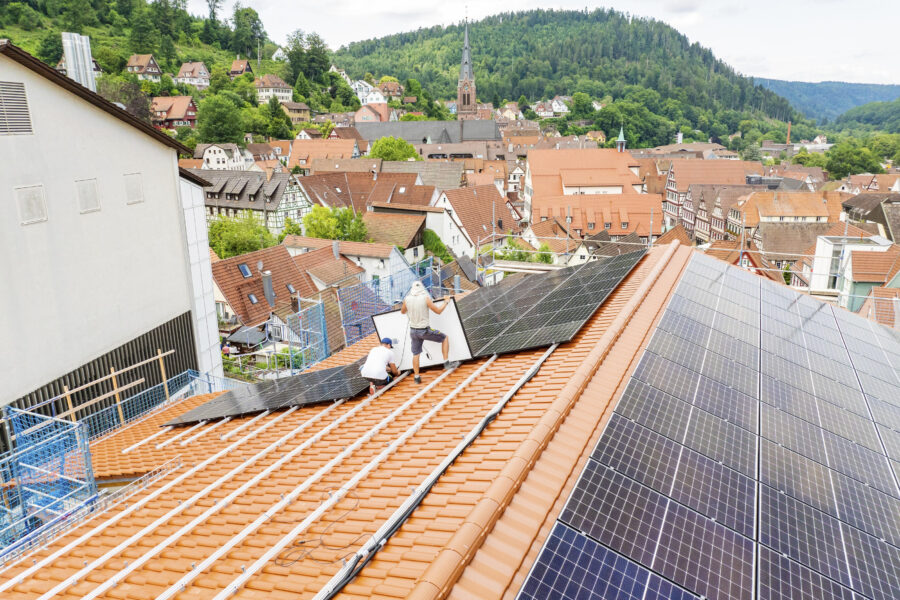 Zwei Personen installieren Solarpaneele auf einem Hausdach mit Blick auf eine Stadt.