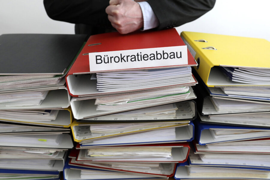 Ordnerstapel mit Schild „Bürokratieabbau“ und einem verschränkten Arm.
