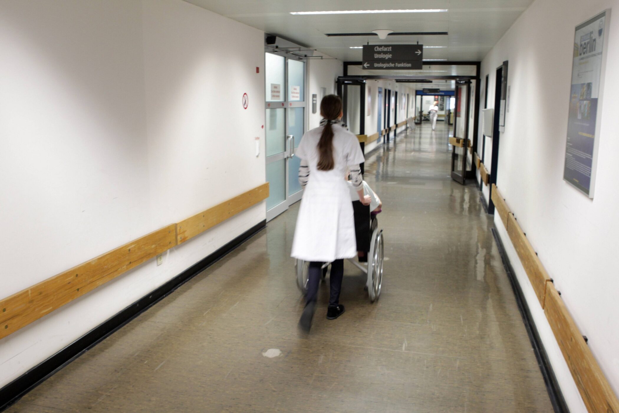 Person schiebt Rollstuhl durch langen Krankenhausflur, Schilder an der Decke.
