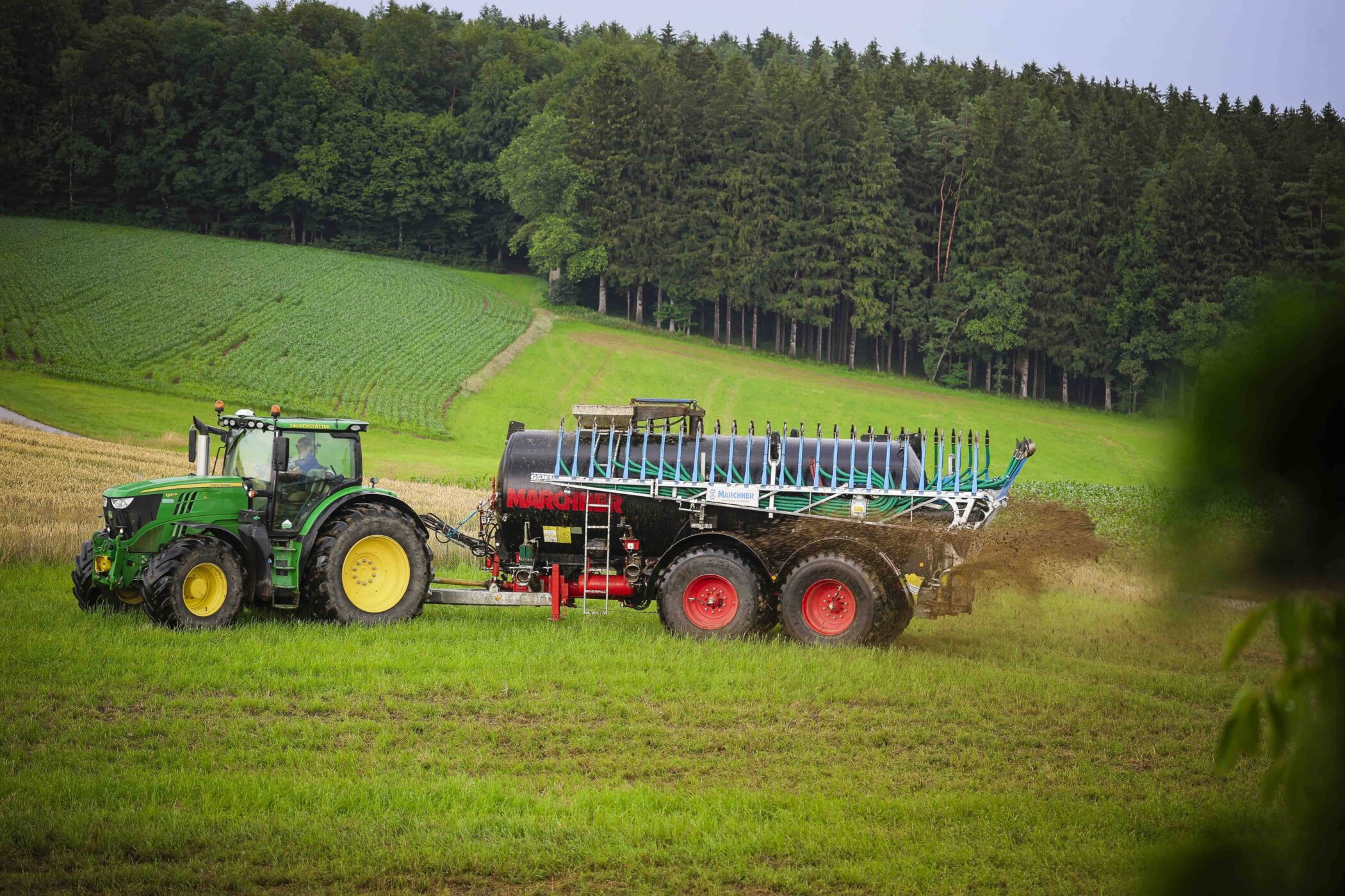 Grüner Traktor mit Güllefass auf einem Feld, Hintergrund mit Wald.