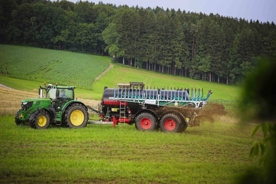 Grüner Traktor mit Güllefass auf einem Feld, Hintergrund mit Wald.