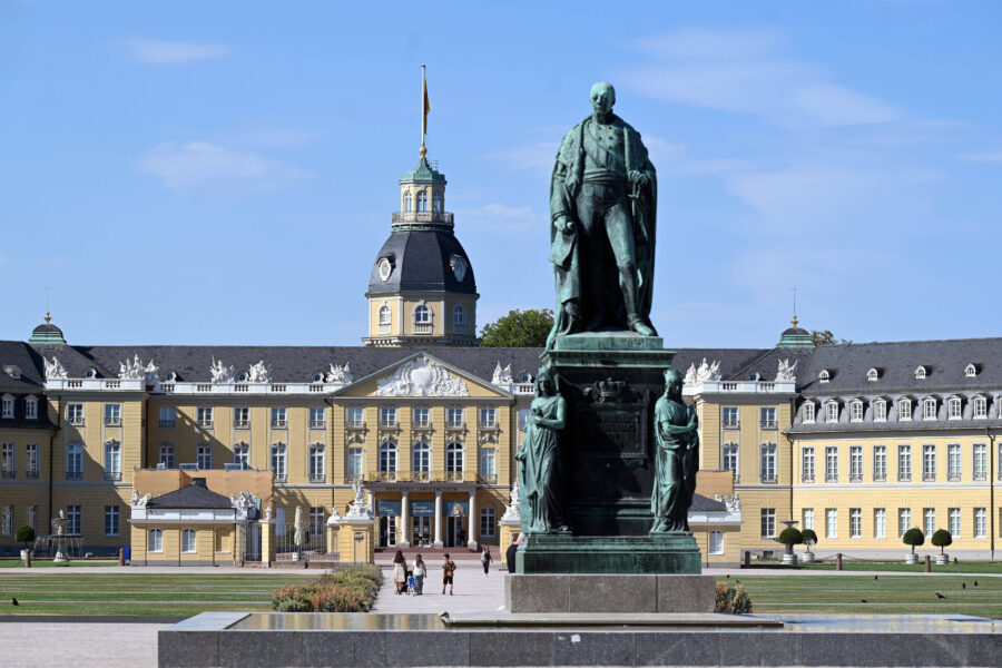 Großes gelbes Gebäude mit Turm; Statue im Vordergrund; Personen spazieren.