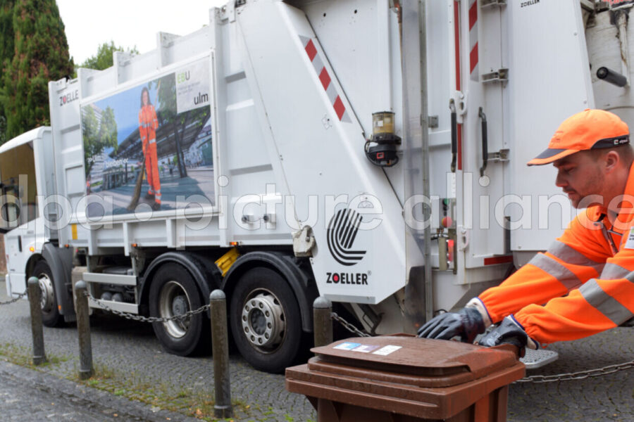 Müllwagen auf Straße, Mann in orangefarbener Arbeitskleidung schiebt Biotonne.