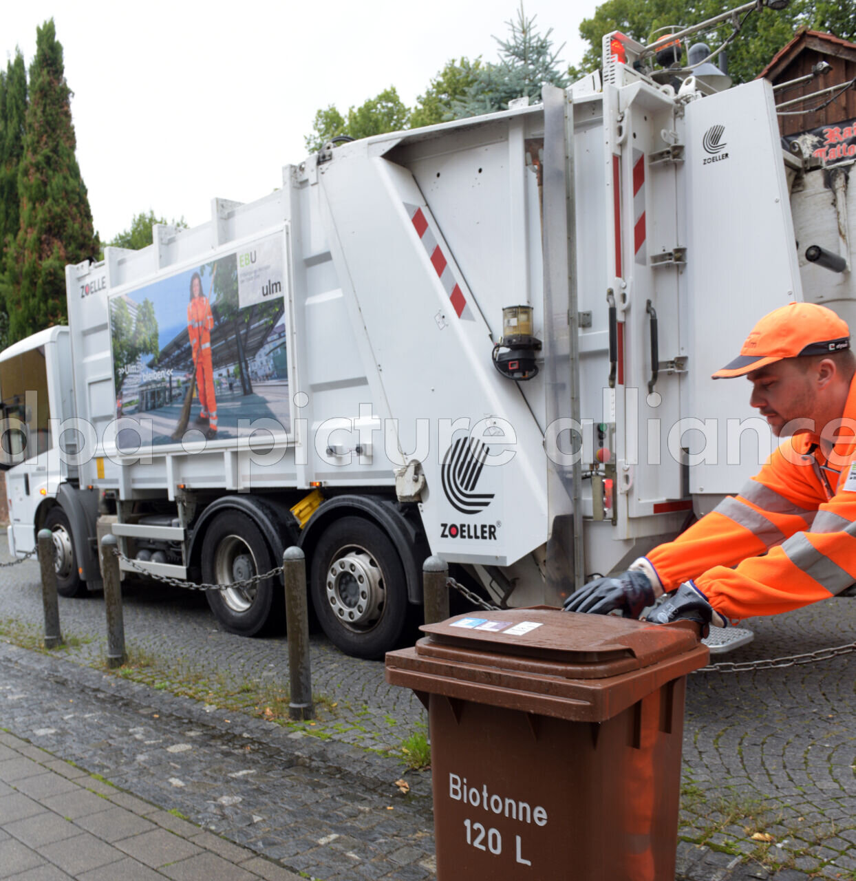 Müllwagen auf Straße, Mann in orangefarbener Arbeitskleidung schiebt Biotonne.