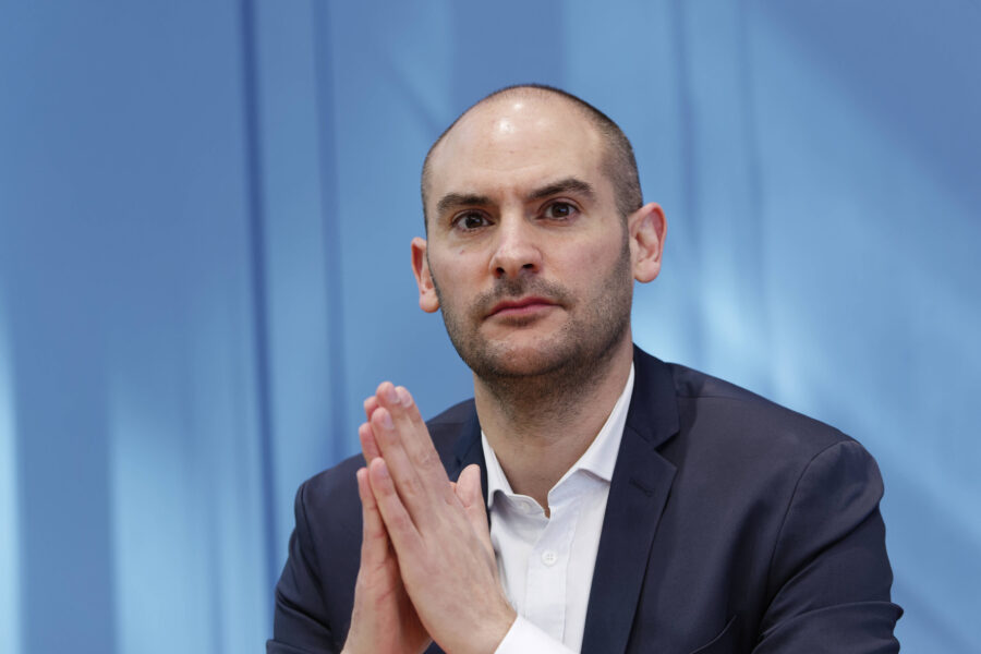 Mann in Anzug mit gefalteten Händen vor blauem Hintergrund.