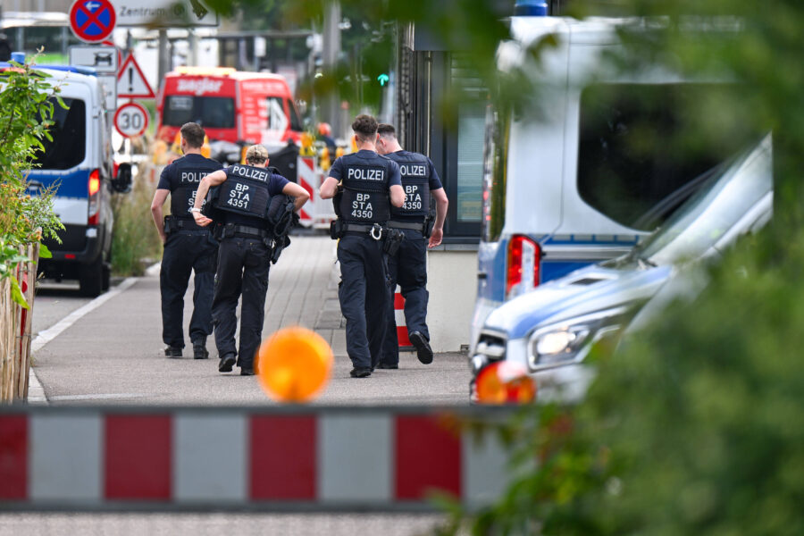Drei Polizisten gehen einen Gehweg entlang, umgeben von Einsatzfahrzeugen.