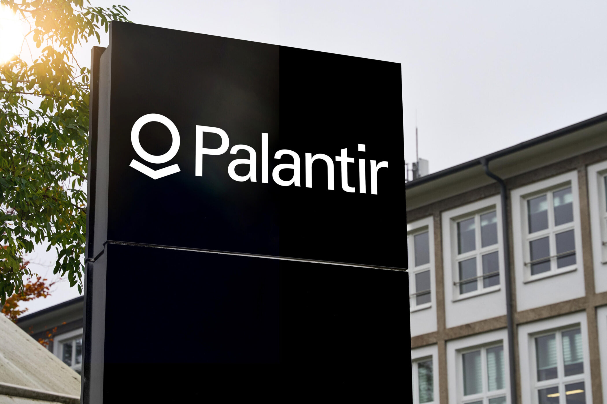 Schwarzes Schild mit weißer Aufschrift "Palantir", im Hintergrund ein Gebäude.