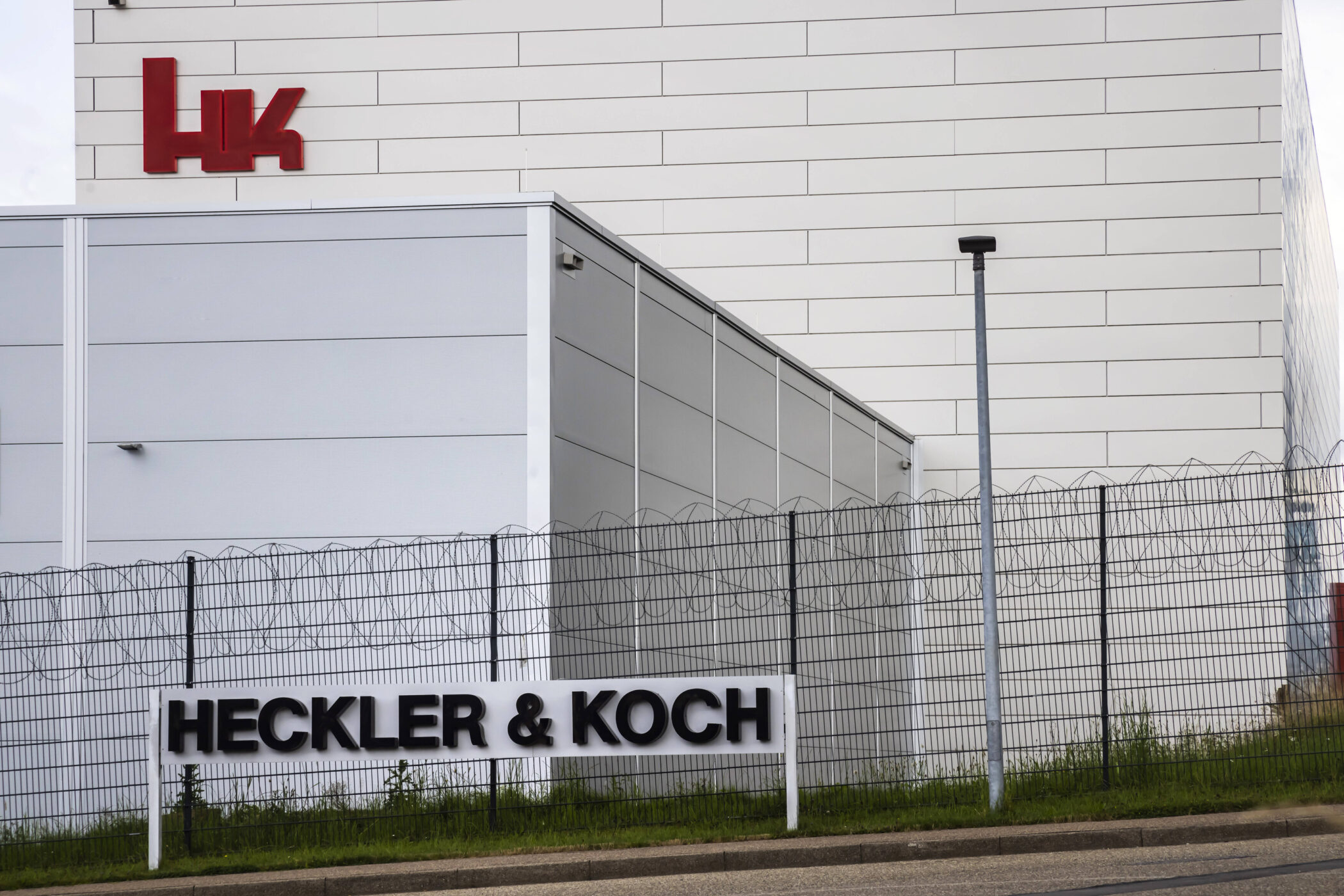 Weißes Gebäude mit rotem "HK"-Logo, Zaun mit "HECKLER & KOCH" Schriftzug.