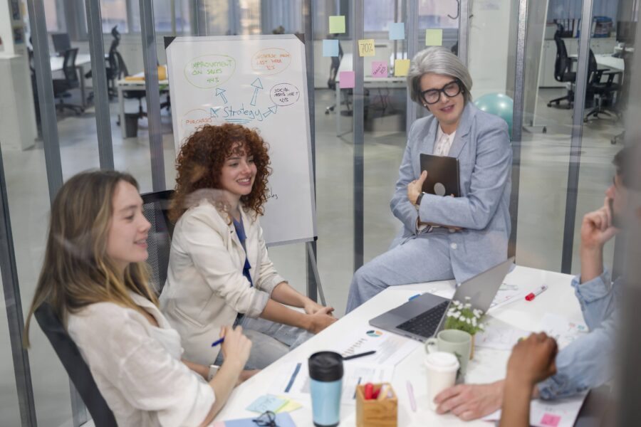 Vier Personen bei einem Meeting; Whiteboard mit Diagrammen im Hintergrund.
