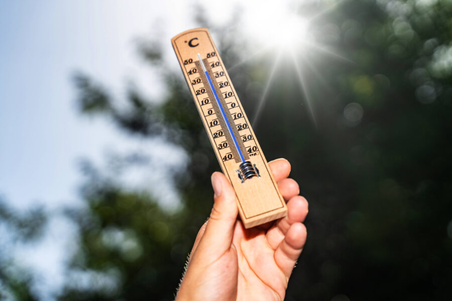 Hand hält Thermometer, zeigt über 30°C, Bäume und Sonnenlicht im Hintergrund.