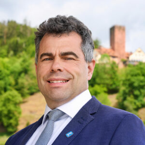Mann in Anzug vor grüner Landschaft mit Turm im Hintergrund.