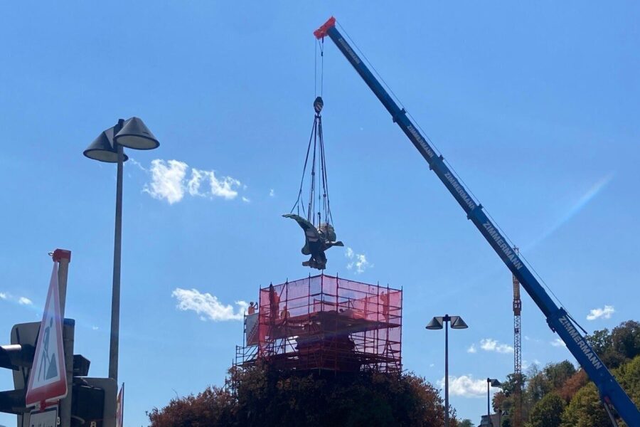 Kran hebt eine Skulptur über ein Gerüst, Ampel und Straßenlaternen im Vordergrund.