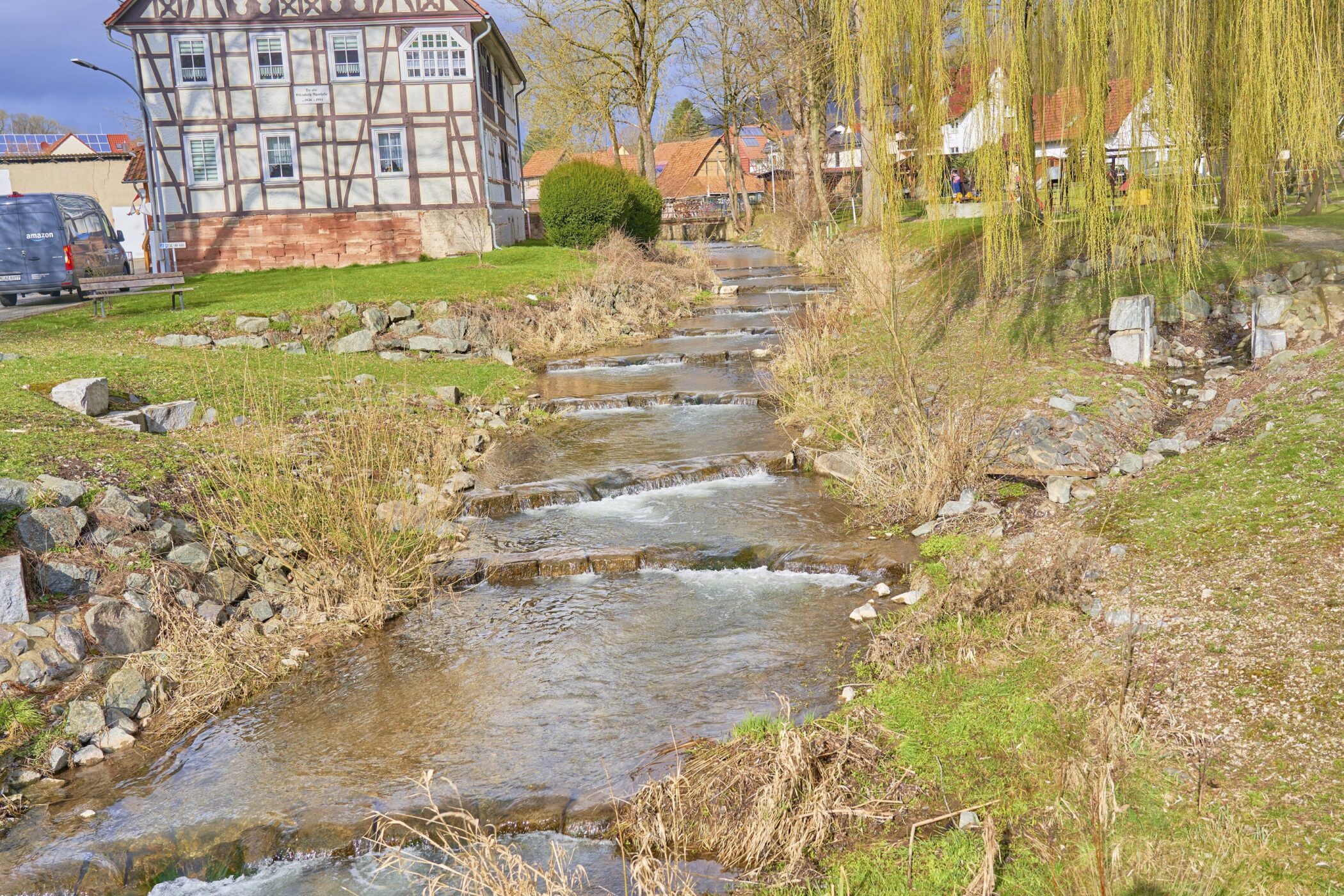 Fachwerkhaus am Bach, Weide hängt über Wiese, Steinmauer am Ufer.