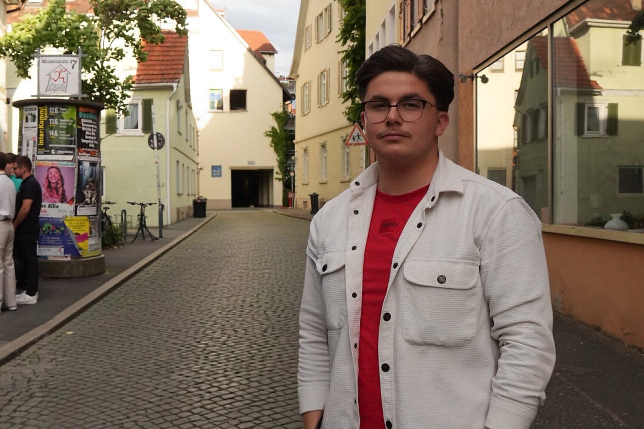 Mann mit Brille und rotem Shirt in gepflasterter Gasse neben Plakatwand.