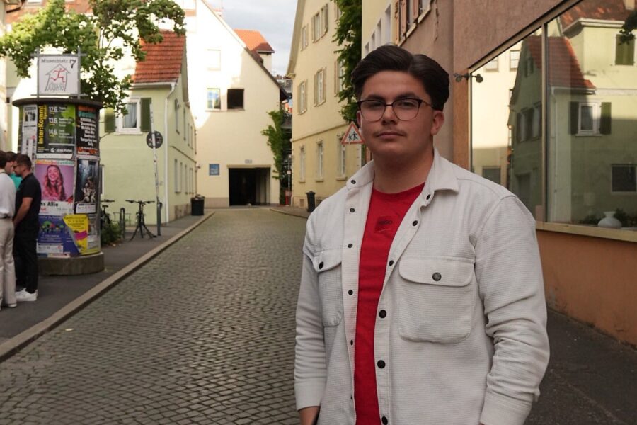 Mann mit Brille und rotem Shirt in gepflasterter Gasse neben Plakatwand.