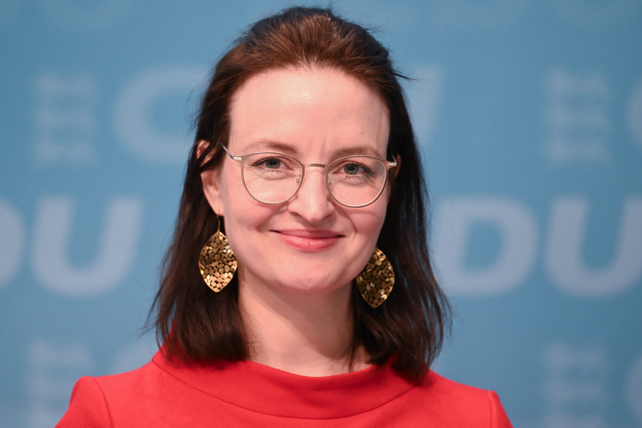 Frau mit Brille und goldenen Ohrringen vor blauem Hintergrund, rotes Oberteil.