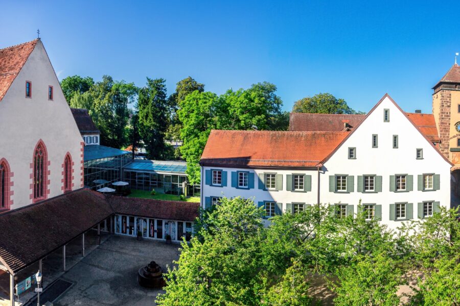 Hof mit historischen Gebäuden, Laube und Turm, umgeben von grünen Bäumen.