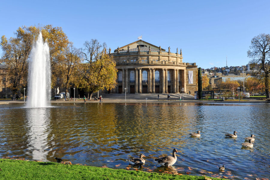 Brunnen und Enten im Teich vor einem Gebäude mit Säulen, Herbstbäume im Hintergrund.