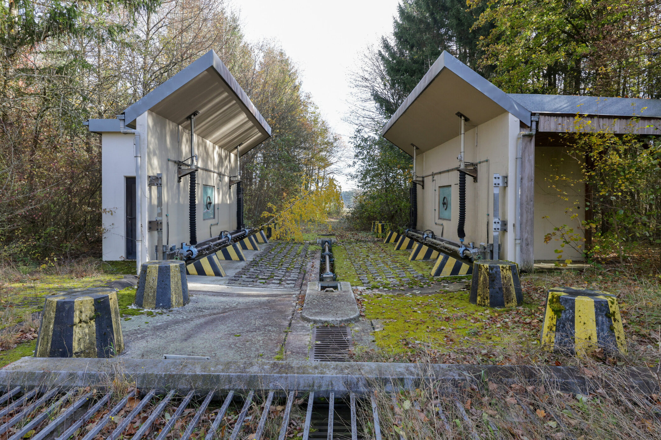 Betongebäude mit offenem Dach, gelb-schwarze Poller, umgeben von Bäumen.