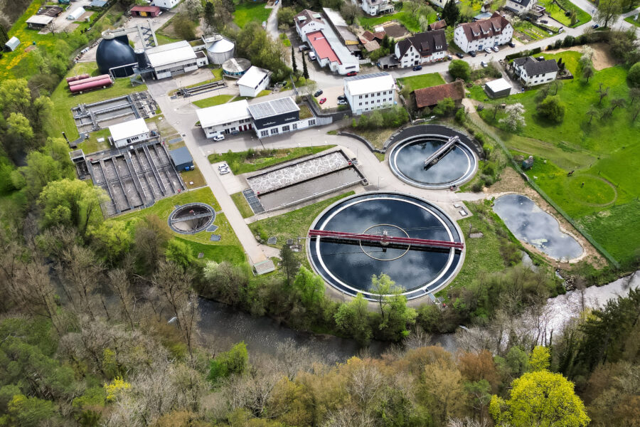 Luftaufnahme einer Kläranlage mit runden und rechteckigen Becken, umgeben von Bäumen und Gebäuden.