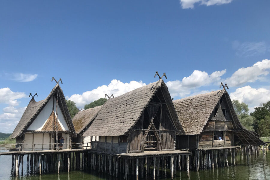 Drei strohgedeckte Pfahlbauten über grünlichem Wasser unter blauem Himmel.