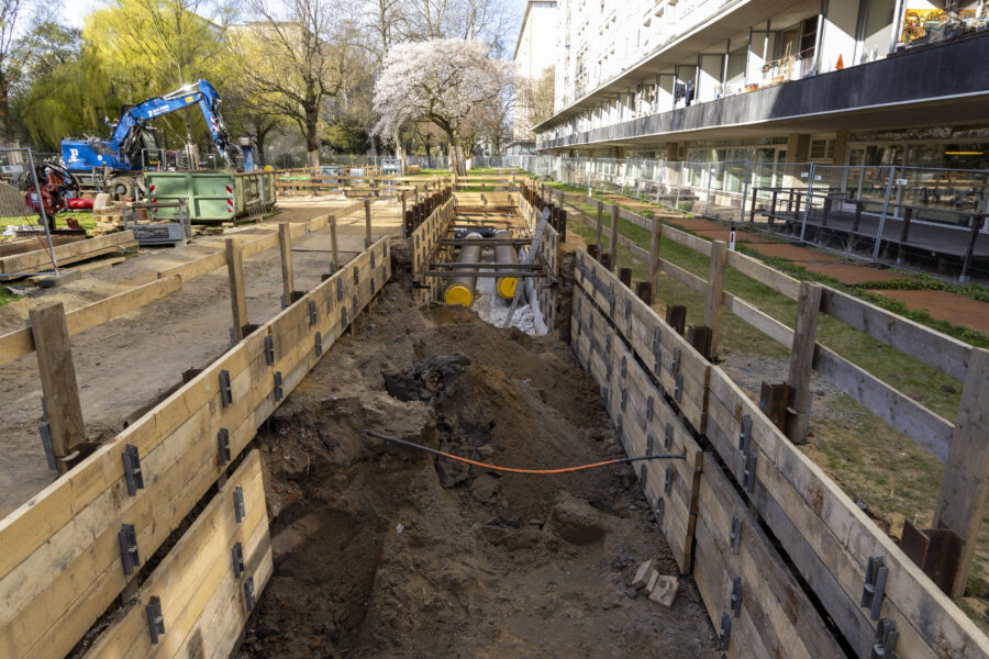 Baugrube mit Bretterverschalung, Bagger und Rohre, Gebäude rechts, Bäume im Hintergrund.
