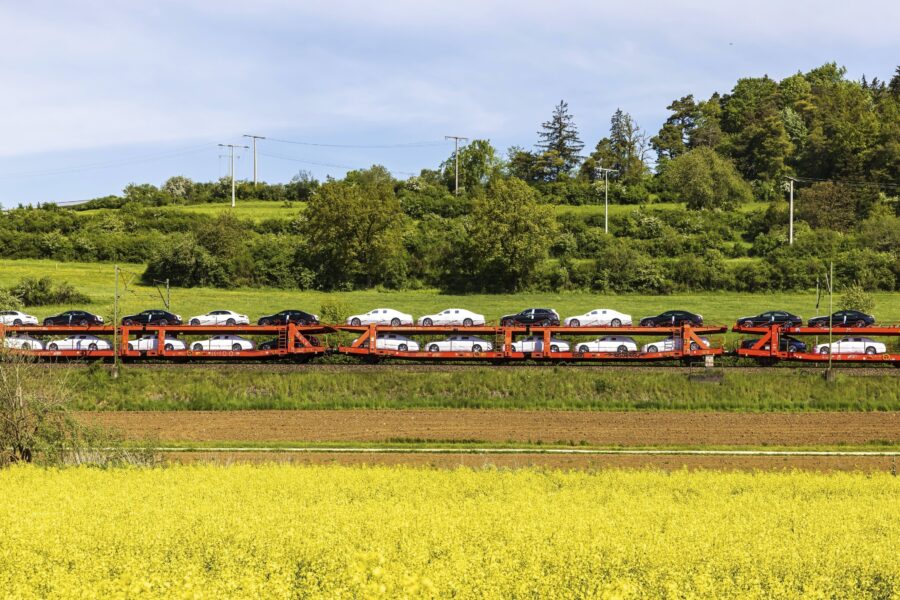 Zug transportiert Autos, fährt durch grüne Landschaft, gelbes Feld im Vordergrund.