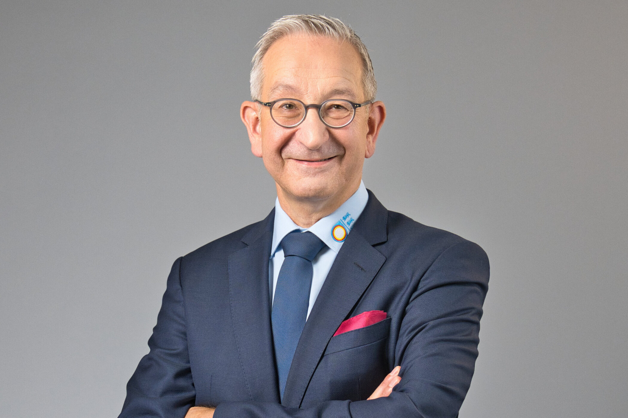 Älterer Mann im Anzug mit Brille, vor neutralem Hintergrund.