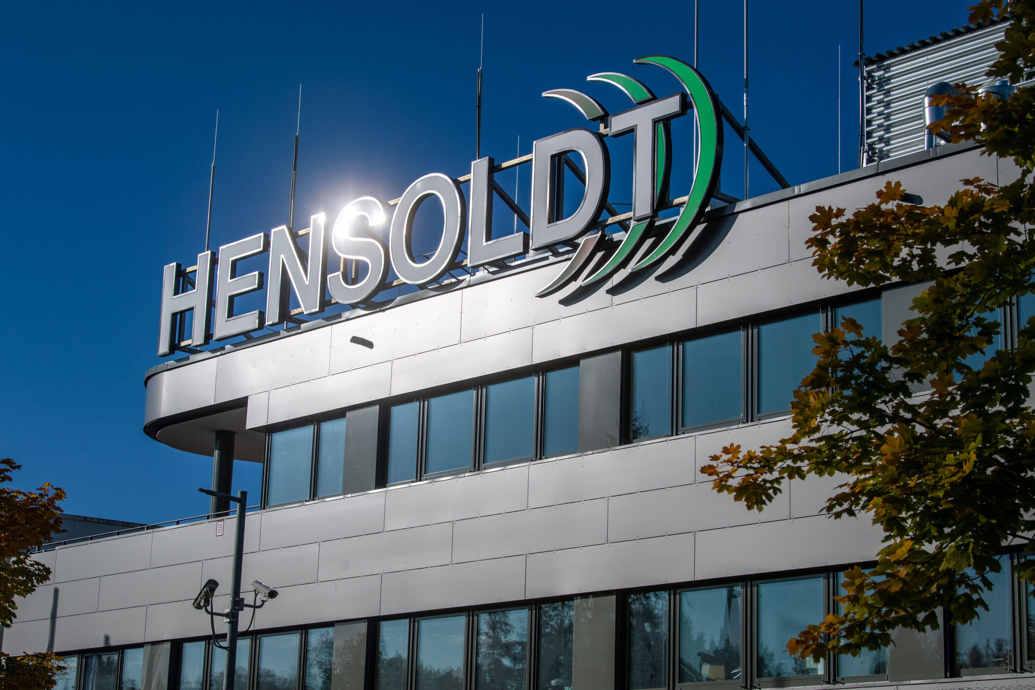 Gebäude mit "Hensoldt" Schriftzug, blauer Himmel, Baum rechts.