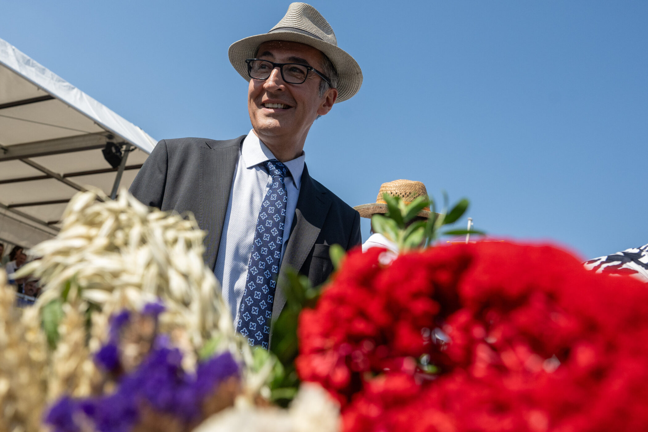 Mann mit Hut und Brille hinter bunten Blumen, blauer Himmel im Hintergrund.