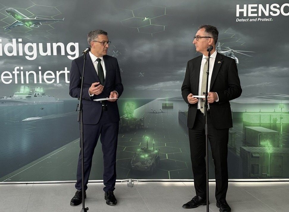 Zwei Männer in Anzügen sprechen vor einem Hintergrund mit Text "Verteidigung definiert".