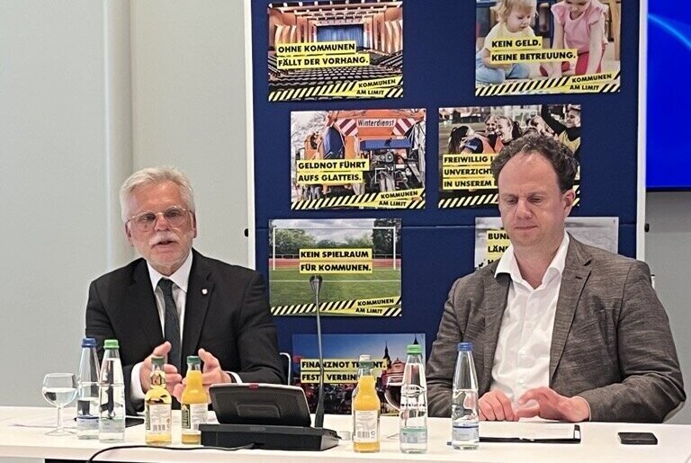 Zwei Männer sitzen an einem Tisch mit Getränken und Mikrofon, Poster im Hintergrund.