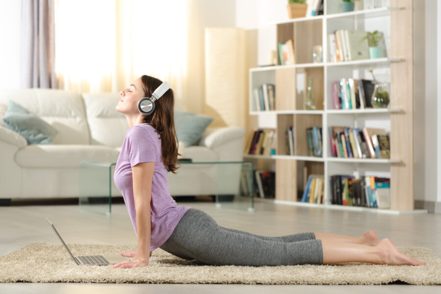 Person macht Yoga mit Kopfhörern, Laptop auf Teppich, Bücherregal im Hintergrund.