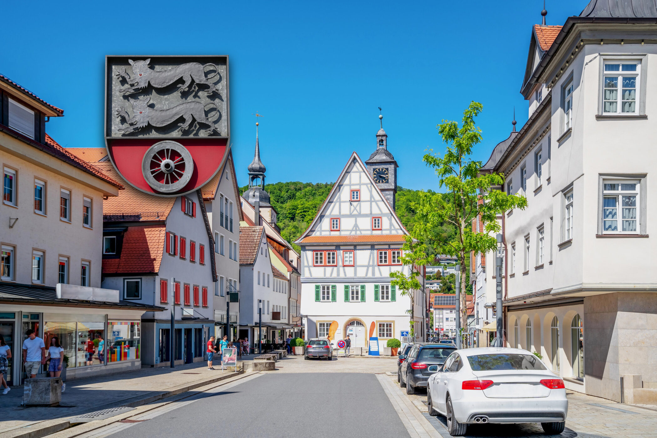 Straße mit Fachwerkhaus, Autos und Fußgängern, blauem Himmel, Wappen oben links.