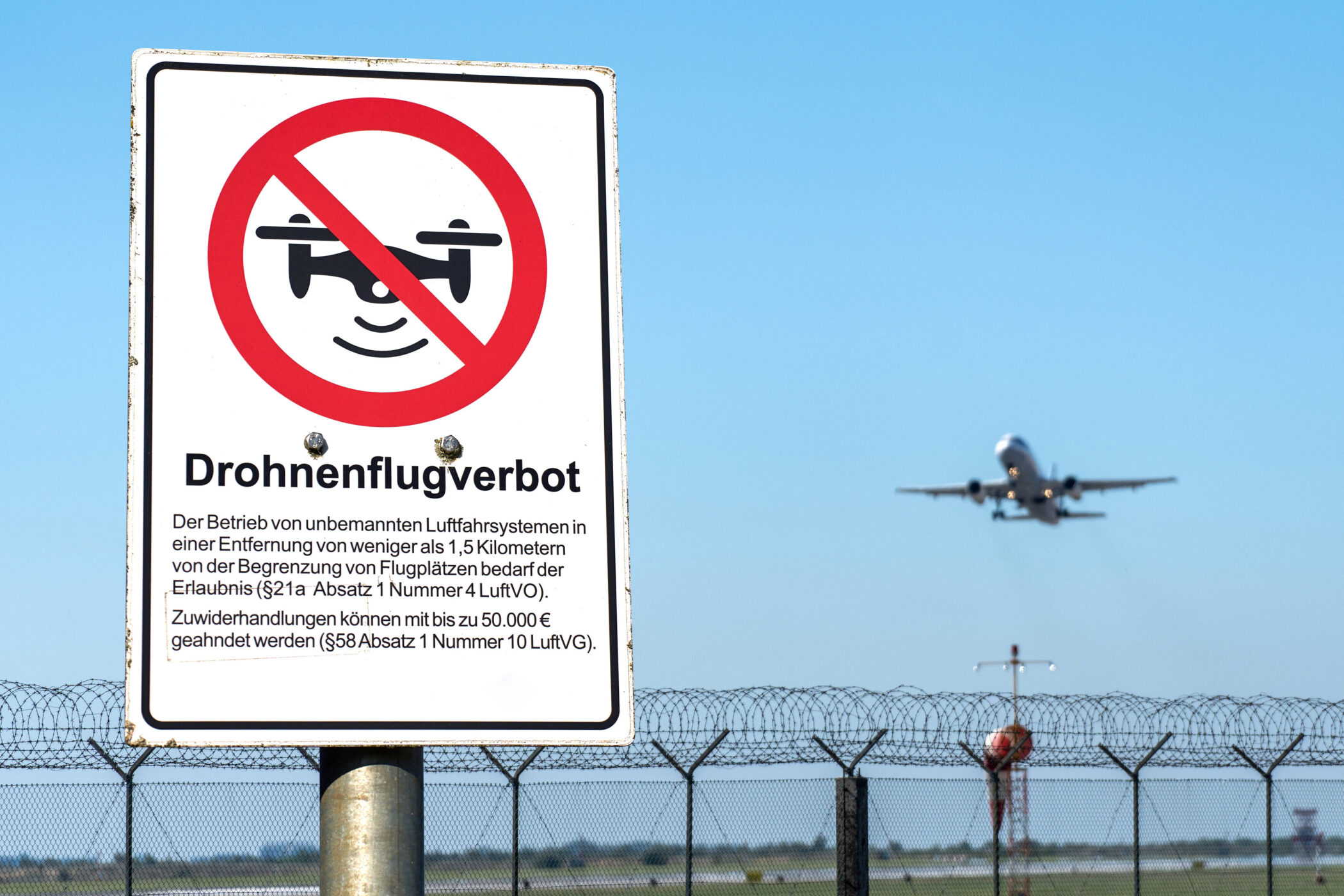 Schild „Drohnenflugverbot“, Flugzeug im Hintergrund, blauer Himmel.