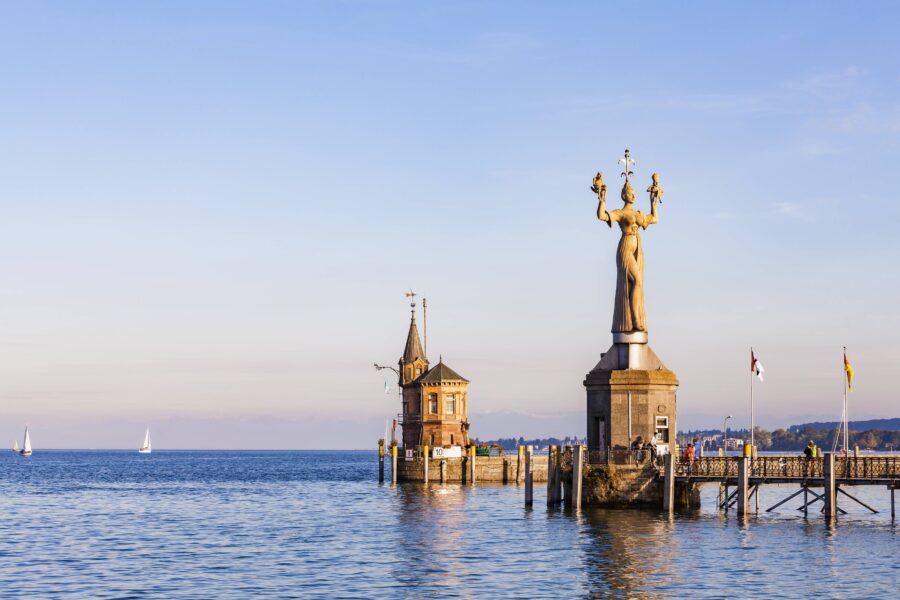 Hafeneinfahrt mit Statue und Turm auf einem Steg, Segelboote auf dem Wasser.