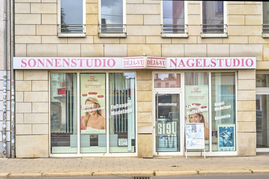Gebäude mit Sonnen- und Nagelstudio, Plakate in Schaufenstern, Eingang rechts.