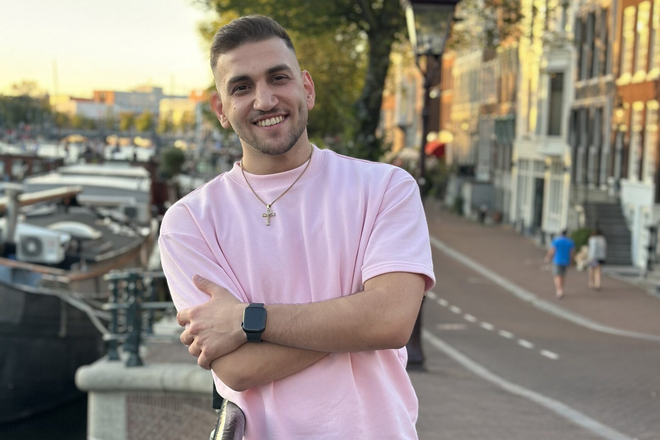 Mann in rosa T-Shirt lächelt, steht vor Kanälen und Gebäuden, zwei Personen im Hintergrund.