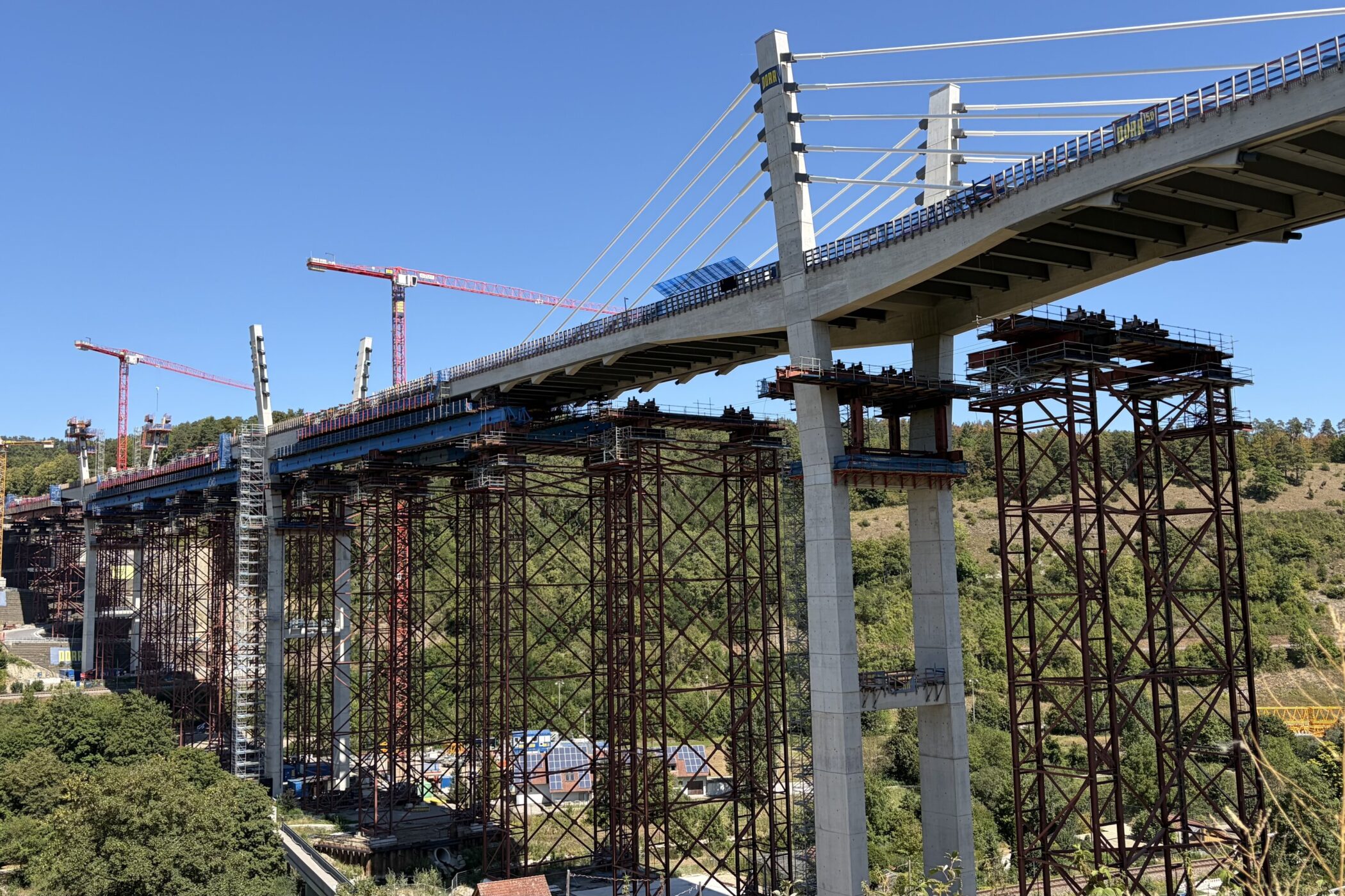 Brücke im Bau, viele Pfeiler und Kräne, bewaldete Hügellandschaft im Hintergrund.