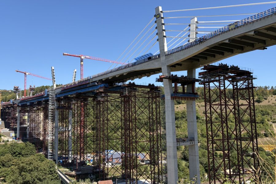 Brücke im Bau, viele Pfeiler und Kräne, bewaldete Hügellandschaft im Hintergrund.