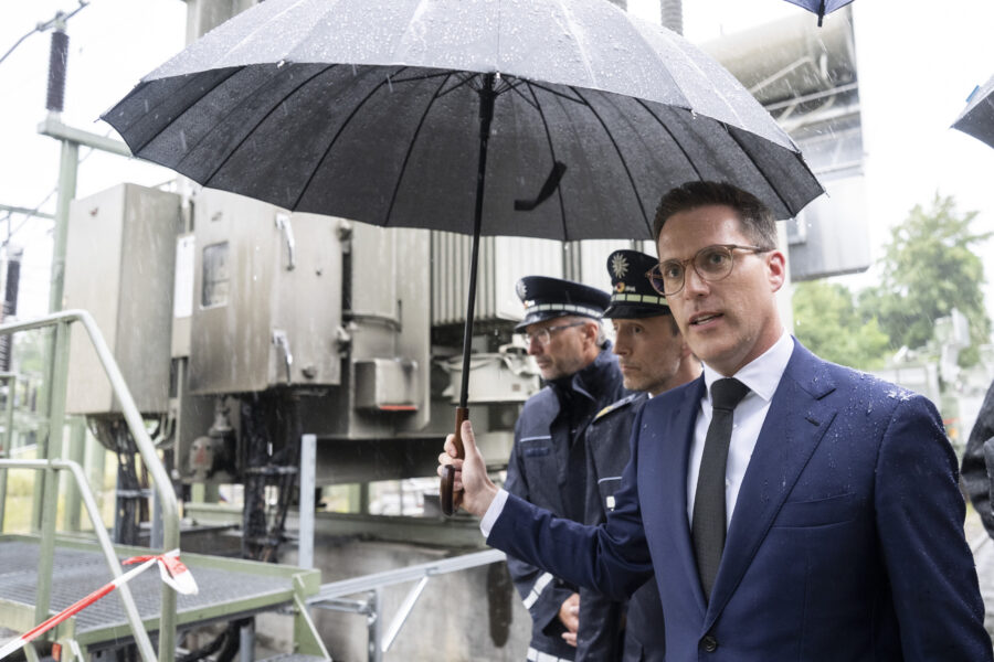 Drei Männer mit Regenschirmen in Anzügen, im Hintergrund industrielle Anlage.