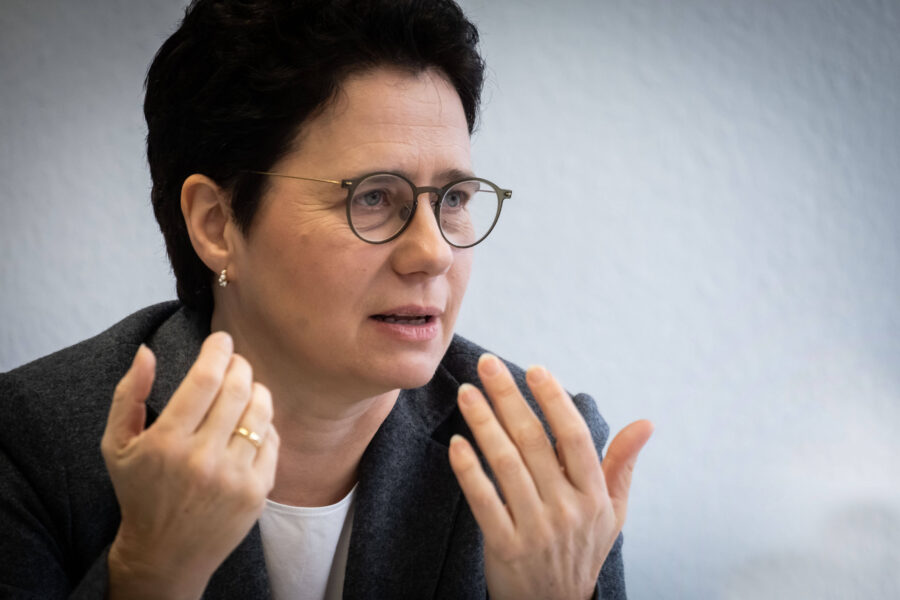 Person mit Brille spricht gestikulierend, grauer Hintergrund.