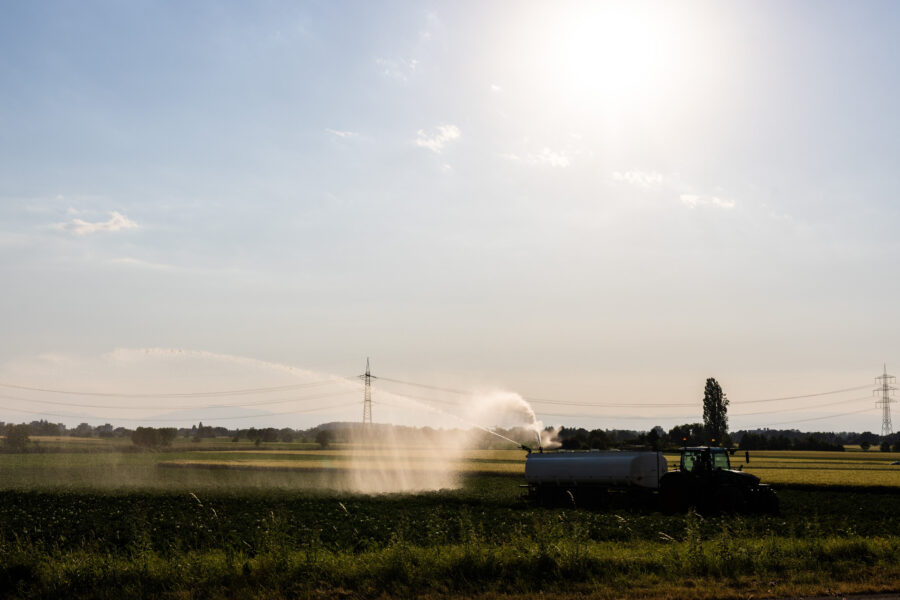 Traktor mit Tank bewässert ein Feld bei Sonnenschein, Stromleitungen im Hintergrund.