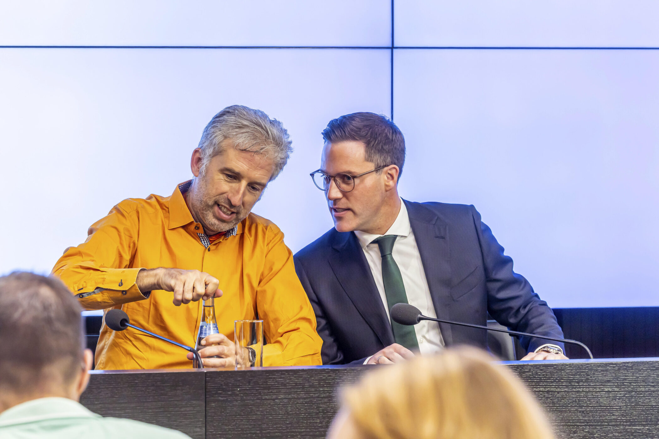 Zwei Männer an einem Tisch mit Mikrofonen, einer öffnet eine Flasche Wasser.
