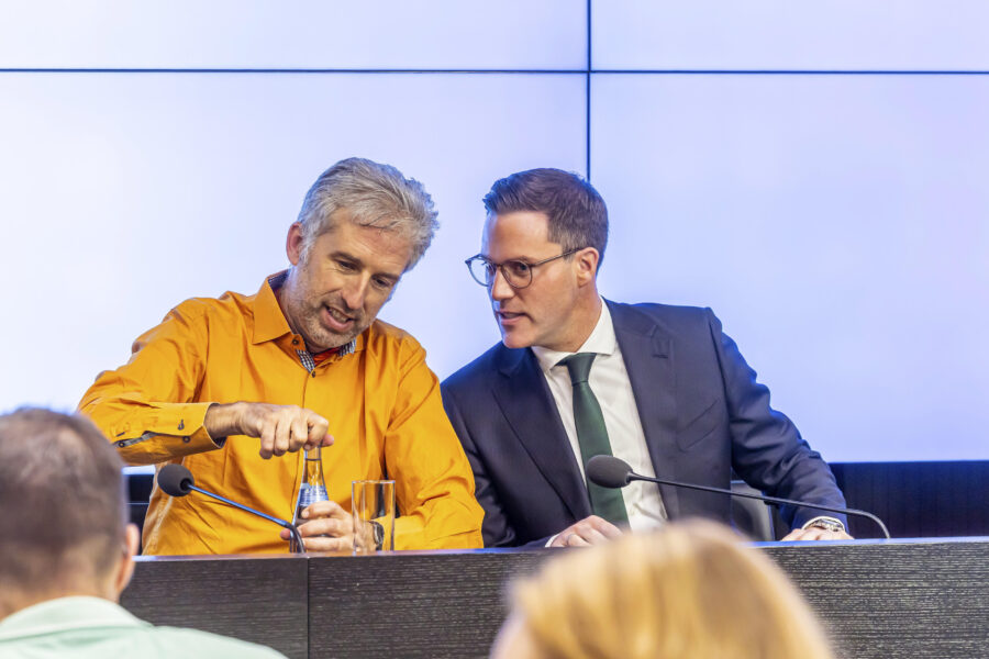Zwei Männer an einem Tisch mit Mikrofonen, einer öffnet eine Flasche Wasser.