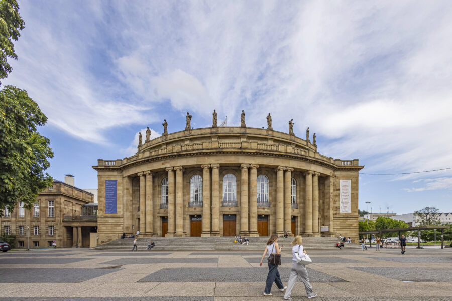 Großes Gebäude mit Säulen und Figuren, Menschen auf Treppen, blauer Himmel.