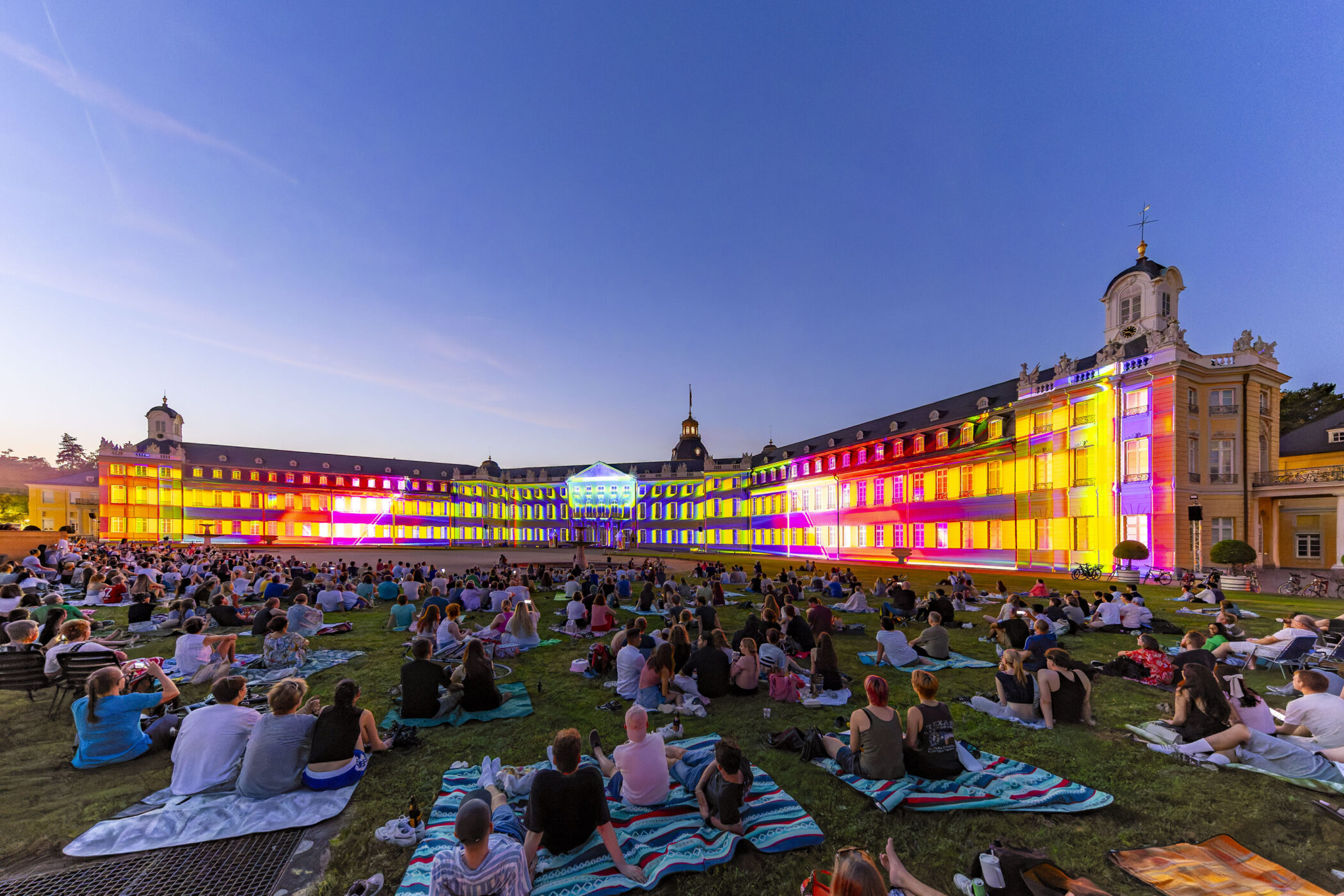 Menschen sitzen auf Wiese vor großem, bunt beleuchteten Gebäude in der Abenddämmerung.