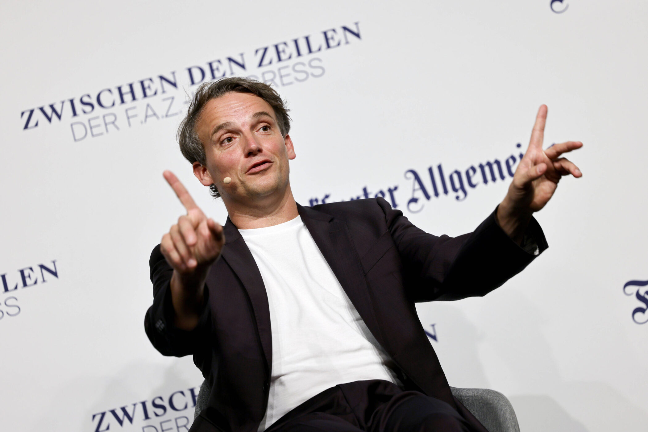 Mann im Anzug sitzt und gestikuliert vor weißer Wand mit Text "Zwischen den Zeilen".
